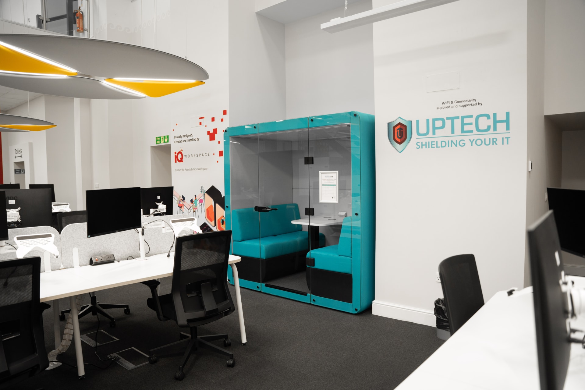 A modern office room with yellow and grey acoustic baffles on the ceiling, a blue acoustic pod, white desks, and black mesh chairs.