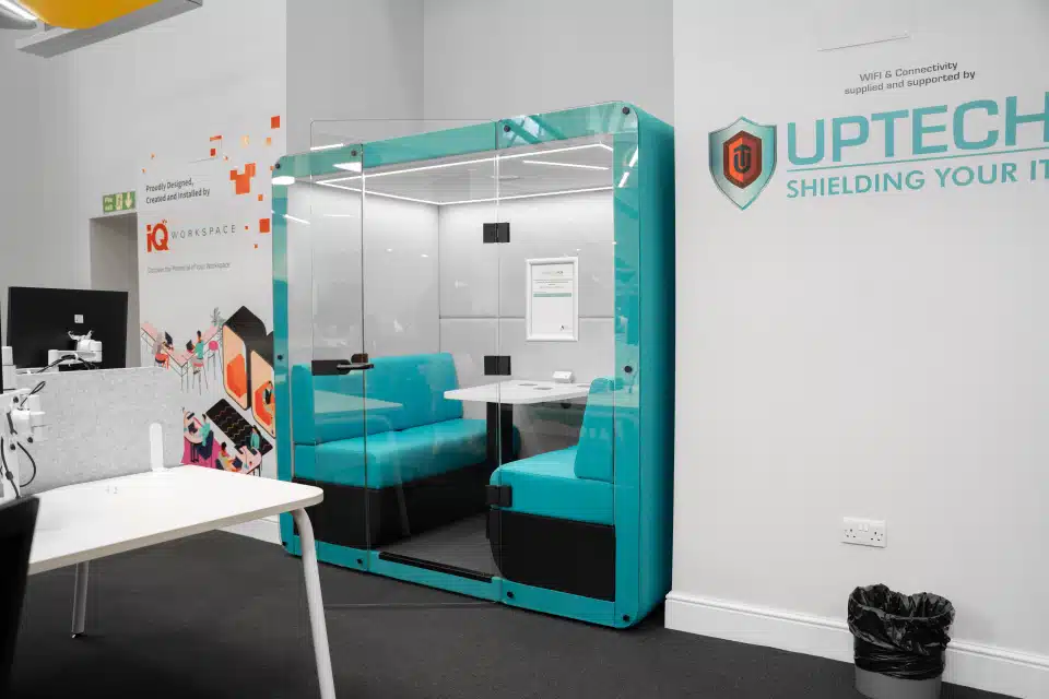 A modern, open-plan office space featuring a blue acoustic pod, white bench desks, and black mesh chairs, with yellow and grey acoustic baffles on the ceiling.