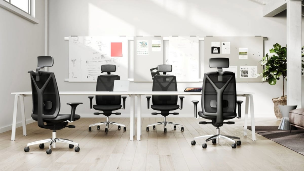 A group of four black mesh chairs placed near white bench desk in an office environment.
