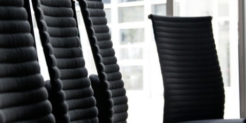 A high-back black leather ergonomic office chair is positioned centrally on a polished concrete floor. It is in a spacious, modern office with large floor-to-ceiling windows displaying a city view.