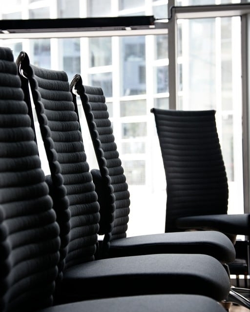 A high-back black leather ergonomic office chair is positioned centrally on a polished concrete floor. It is in a spacious, modern office with large floor-to-ceiling windows displaying a city view.