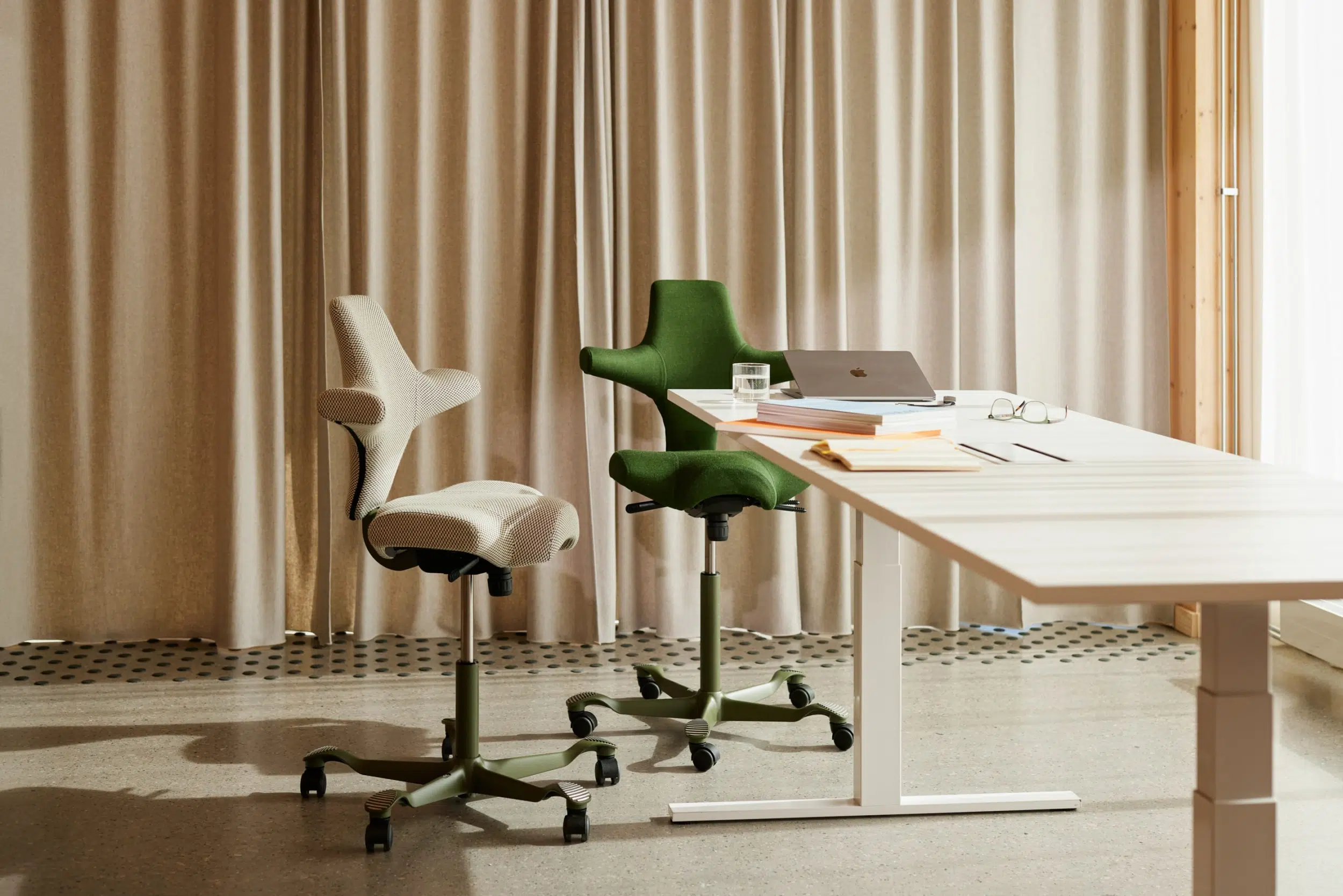 Two HAG Capisco ergonomic saddle chairs in green and cream beside a white standing desk.