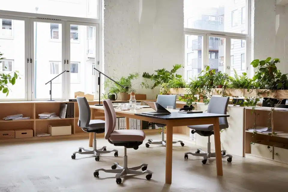 Multiple HÅG Creed ergonomic task chairs (pink and grey) surrounding a collaborative wooden workspace filled with plants.