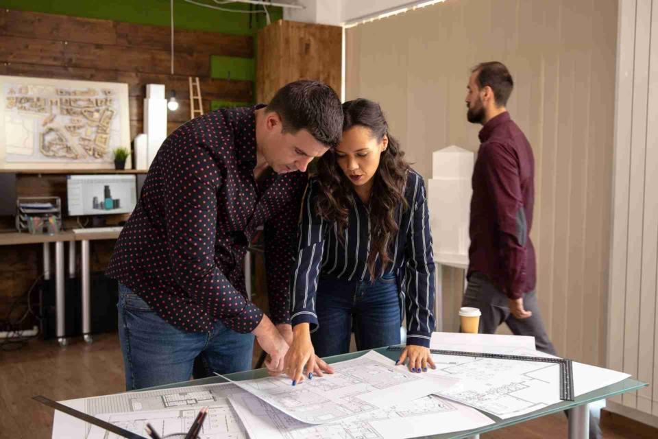 Two architects, a man and a woman, are standing at a modern office table reviewing a project together.