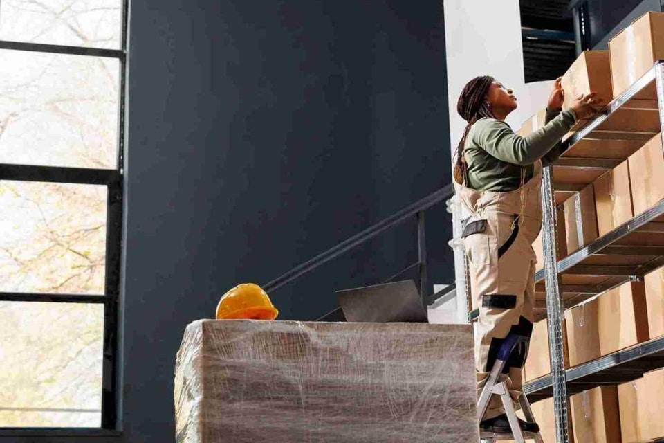 Stockroom worker on a ladder in a large, organised warehouse, holding a cardboard box as they prepare customer orders and check packages for shipping.