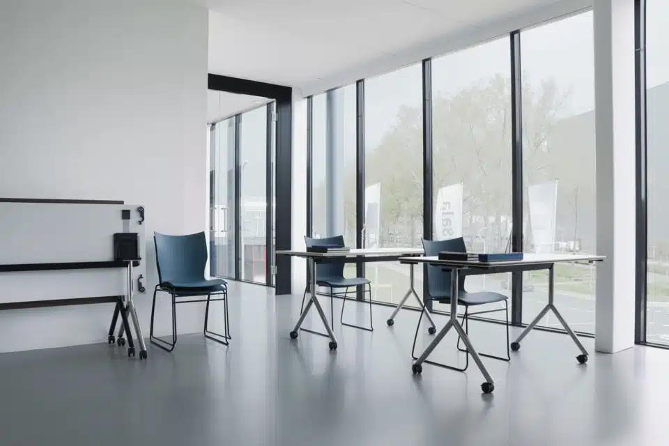 Row of blue plastic canteen chairs and modular wooden school-style desks arranged in a linear office layout.