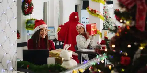 A female in festive attire surprises his coworkers with Christmas gifts.