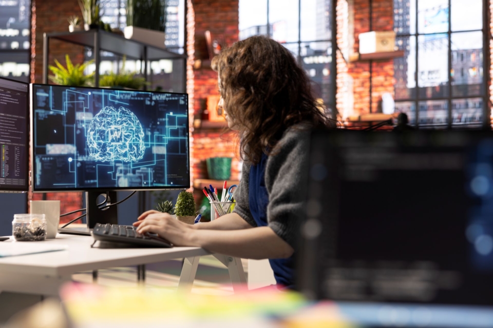 An office worker typing at a desk with a monitor displaying a glowing blue digital brain graphic.