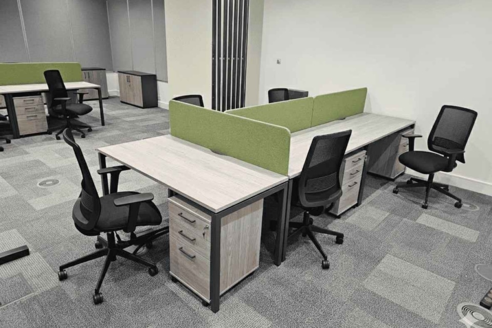 This modern office workspace features back-to-back light-wood desks with green privacy screens, black-mesh ergonomic chairs, and grey carpet tiles.
