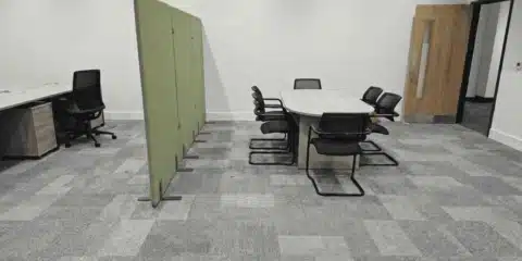 Modern open-plan office interior featuring a light wood meeting table with black chairs, a green acoustic room divider, and a grey carpeted floor with integrated power outlets.