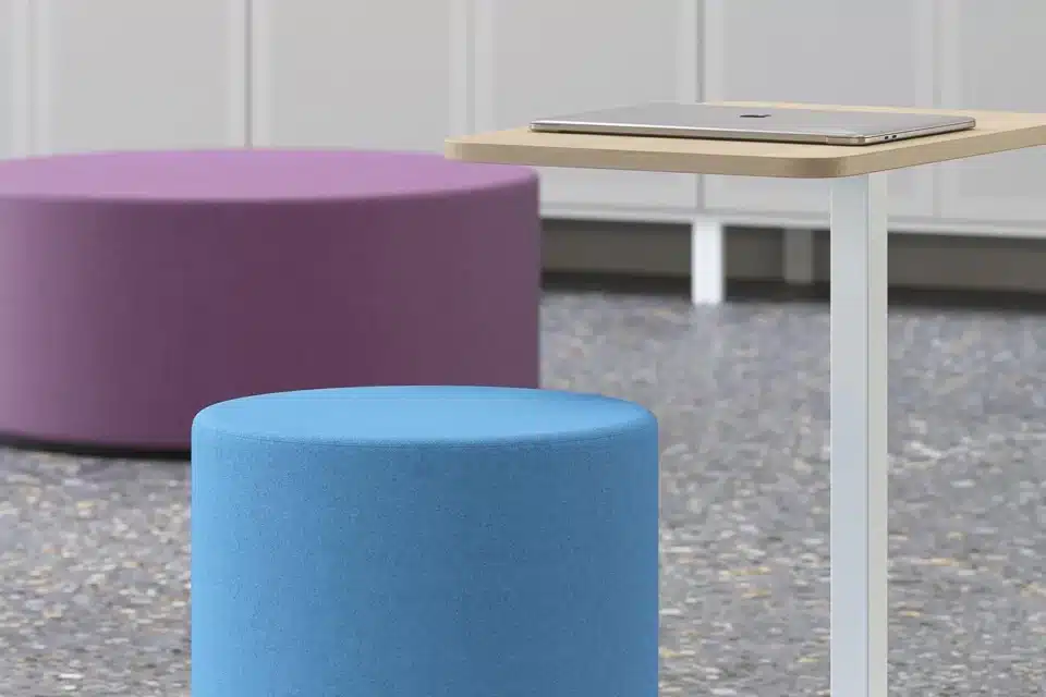 Close-up of a light wood MOBI tabletop with a laptop on it, positioned next to a bright blue and a purple circular pouffe.