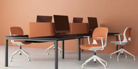 Ace swivel office chairs with orange cushions and white frames are positioned at a long black desk in a modern peach-colored workspace.