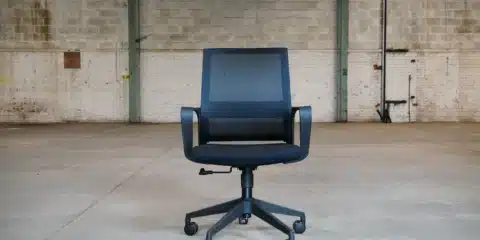 Front view of a black Oslo mesh office chair with integrated armrests and a five-star wheeled base, set against a large industrial warehouse background.