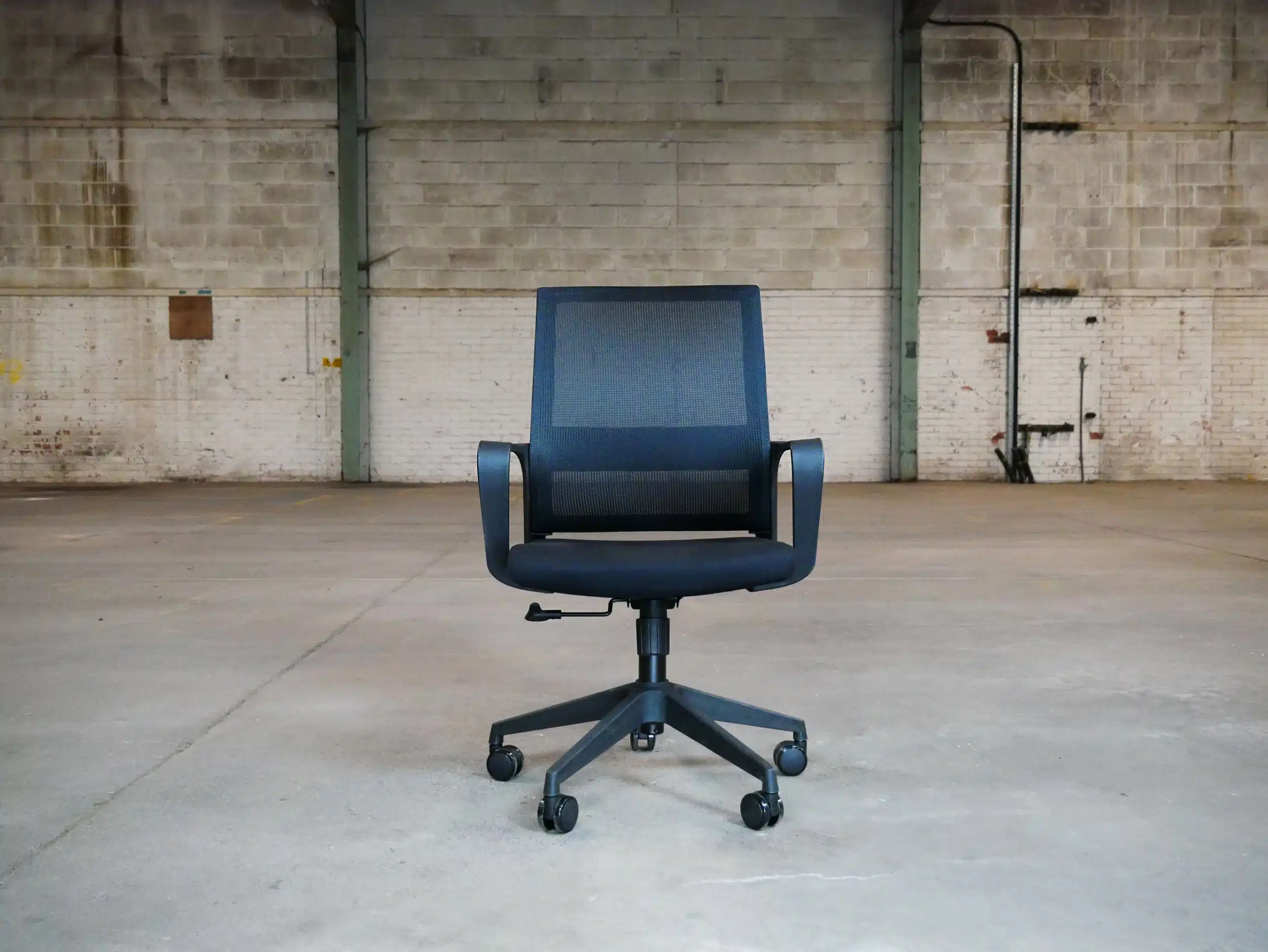 Front view of a black Oslo mesh office chair with integrated armrests and a five-star wheeled base, set against a large industrial warehouse background.