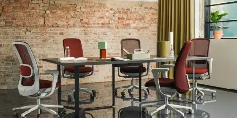 Five HÅG SoFi mesh ergonomic chairs in deep red and light grey arranged around a dark minimalist conference table in an industrial-style office with an exposed brick wall.