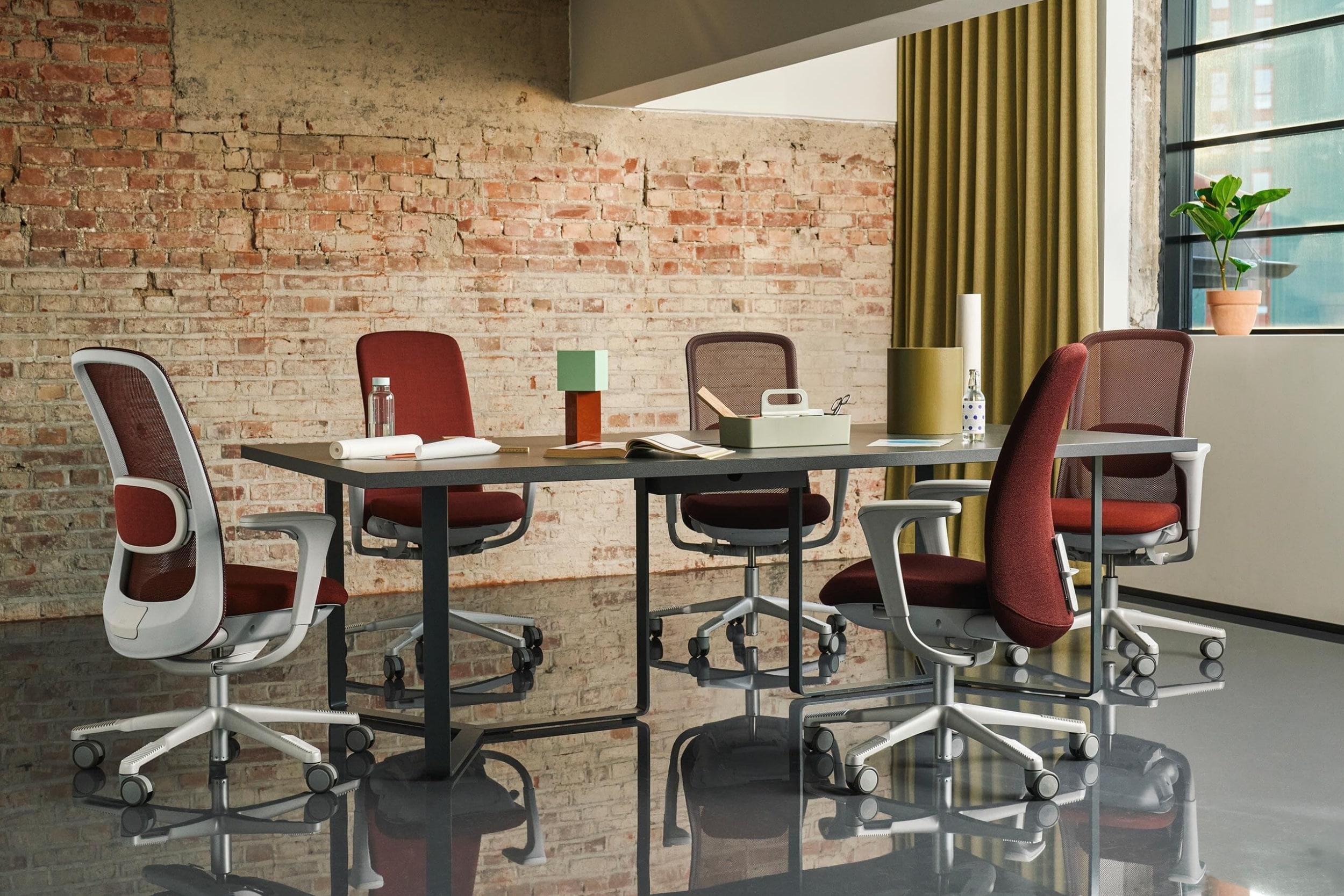 Five HÅG SoFi mesh ergonomic chairs in deep red and light grey arranged around a dark minimalist conference table in an industrial-style office with an exposed brick wall.