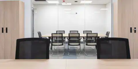 A modern office setting viewed through a glass partition, featuring several black mesh-back "Madrid" cantilever chairs arranged around a light wood conference table.