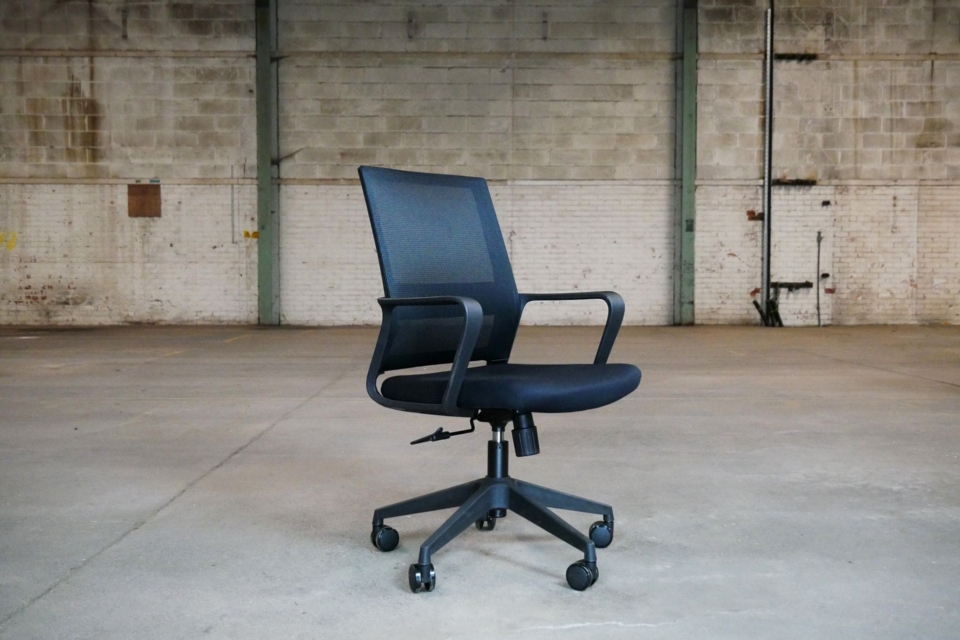 A black mesh swivel office chair with armrests, positioned in the center of a spacious, industrial warehouse with concrete floors and brick walls.