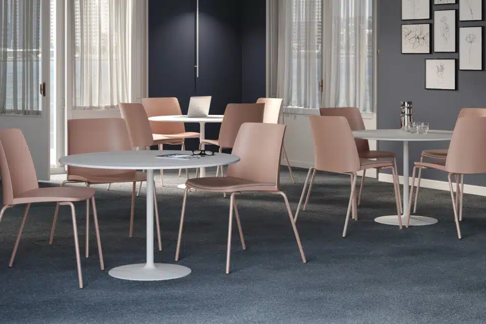 A group of blush pink Seattle meeting chairs with matching metal legs arranged around white circular pedestal tables in a dark grey, modern office lounge.