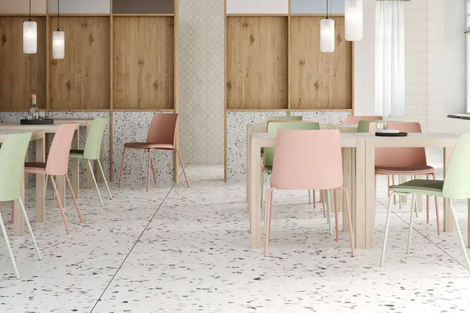 A bright canteen area featuring Seattle chairs in sage green, soft pink, and terracotta, arranged around light wooden tables on a terrazzo floor.