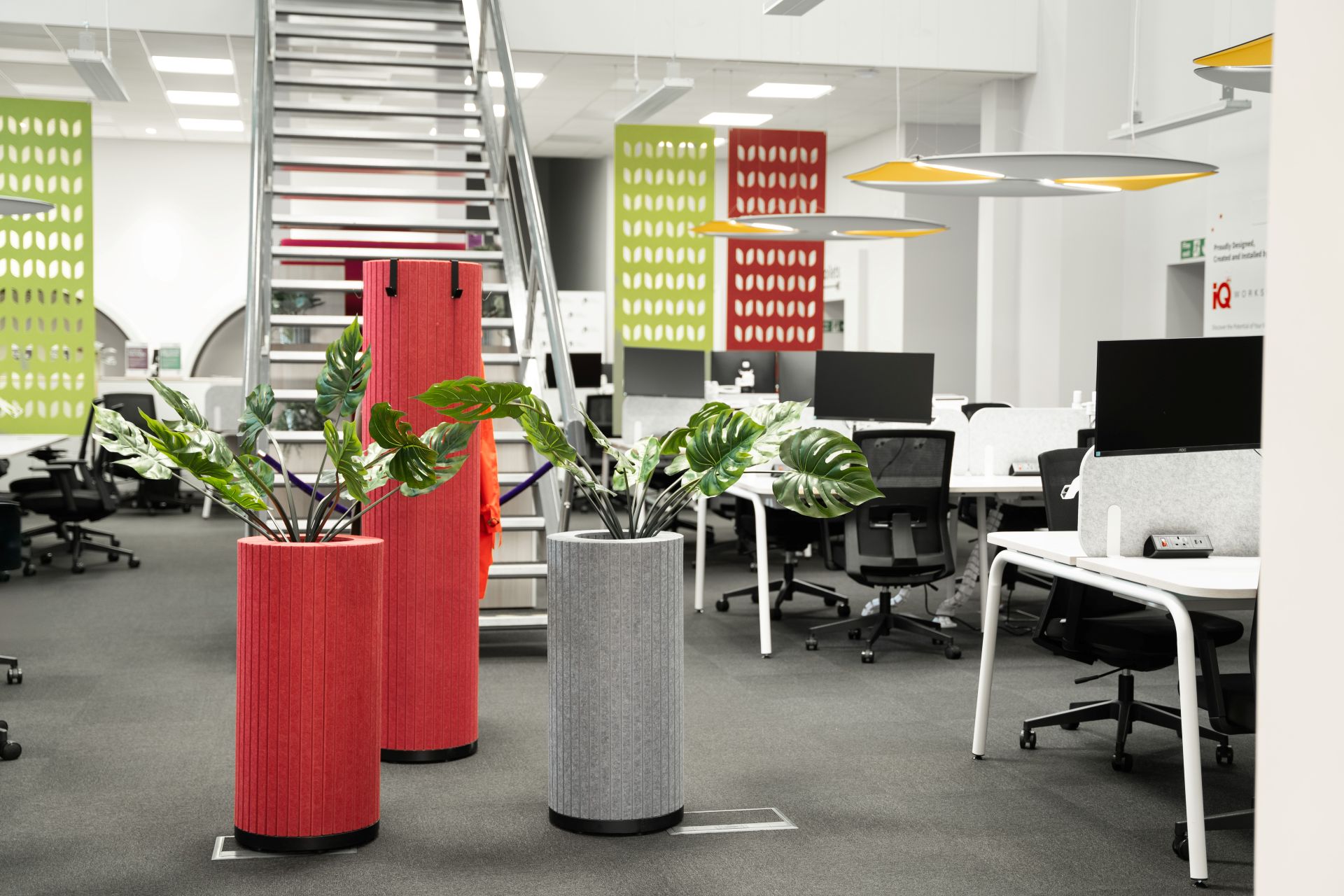 Modern open-plan office interior featuring acoustic felt planters in red and grey, colorful hanging sound baffles, ergonomic white desks, and industrial metal stairs.