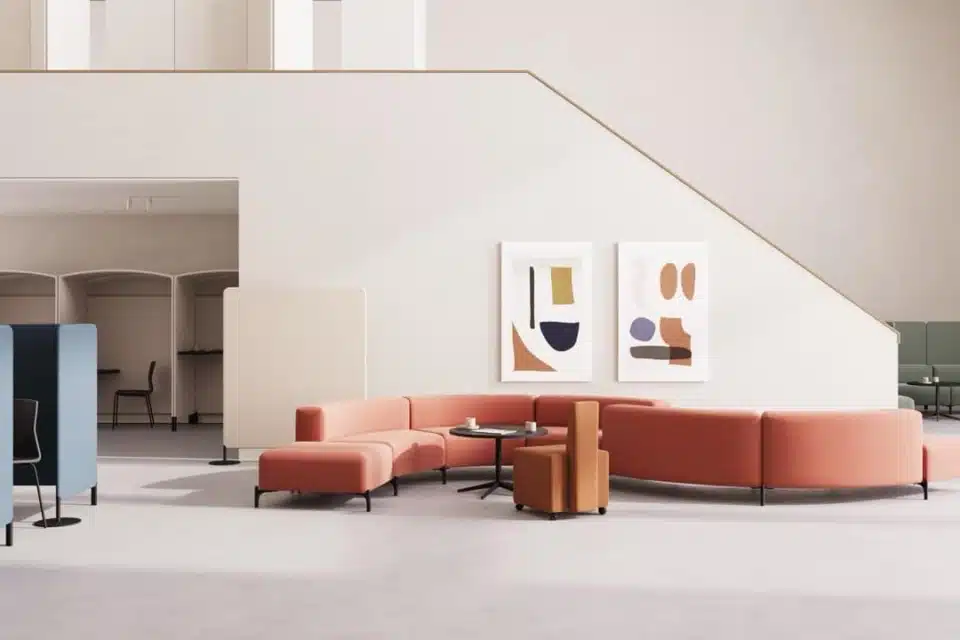A modern, open-plan office lounge featuring a modular, curved terracotta-colored sofa arranged in a semi-circle around a black coffee table. The space is minimalist, with high ceilings, neutral white walls, abstract art, and blue acoustic privacy screens.