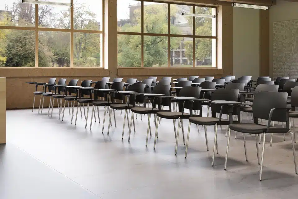 Modern lecture theatre featuring rows of dark grey chairs with writing tablets, expansive light wood windows showing autumn trees, and neutral colours suitable for an iQ Workspace environment.