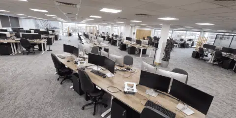 A wide-angle view of a large, empty open-plan office featuring rows of wooden desks with computer monitors, black chairs, and daylight shining through large windows.