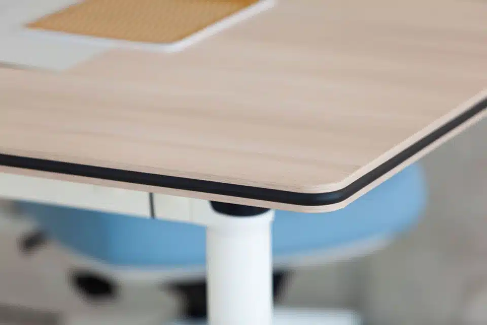 Close-up of the Talent desk surface, showcasing the light wood melamine finish, rounded corners, and protective rubber edges for safety.