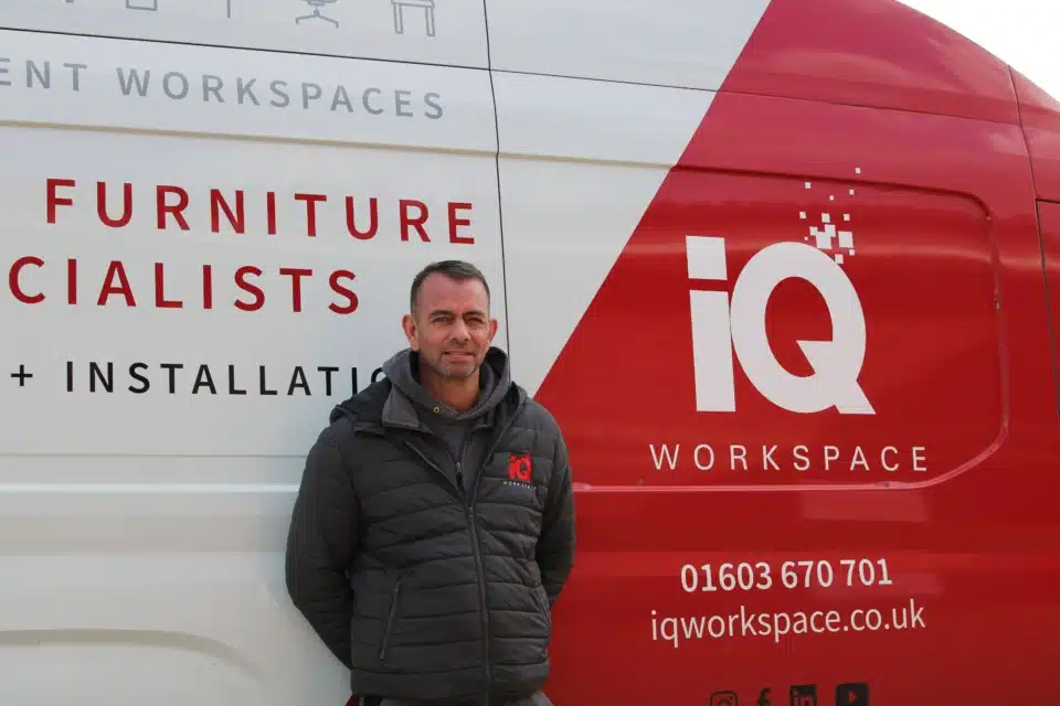 A smiling iQ Workspace installer in a branded puffer jacket standing proudly in front of a white and red company delivery van.