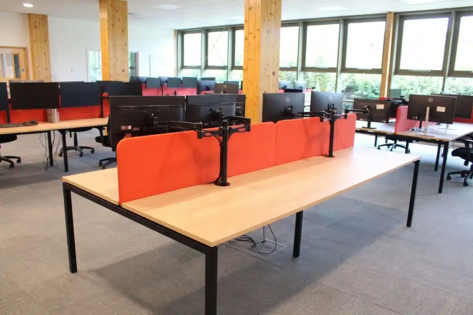 Sunlit office interior featuring modern workstations with red dividers and black monitor stands.