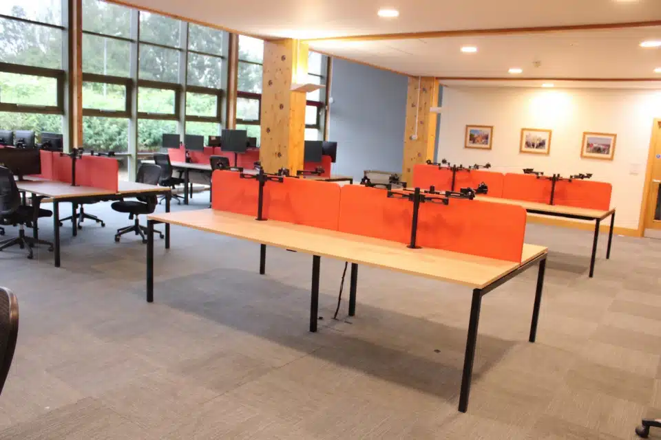 Modern commercial office furniture setup showing desks with red acoustic panels and large wooden pillars.