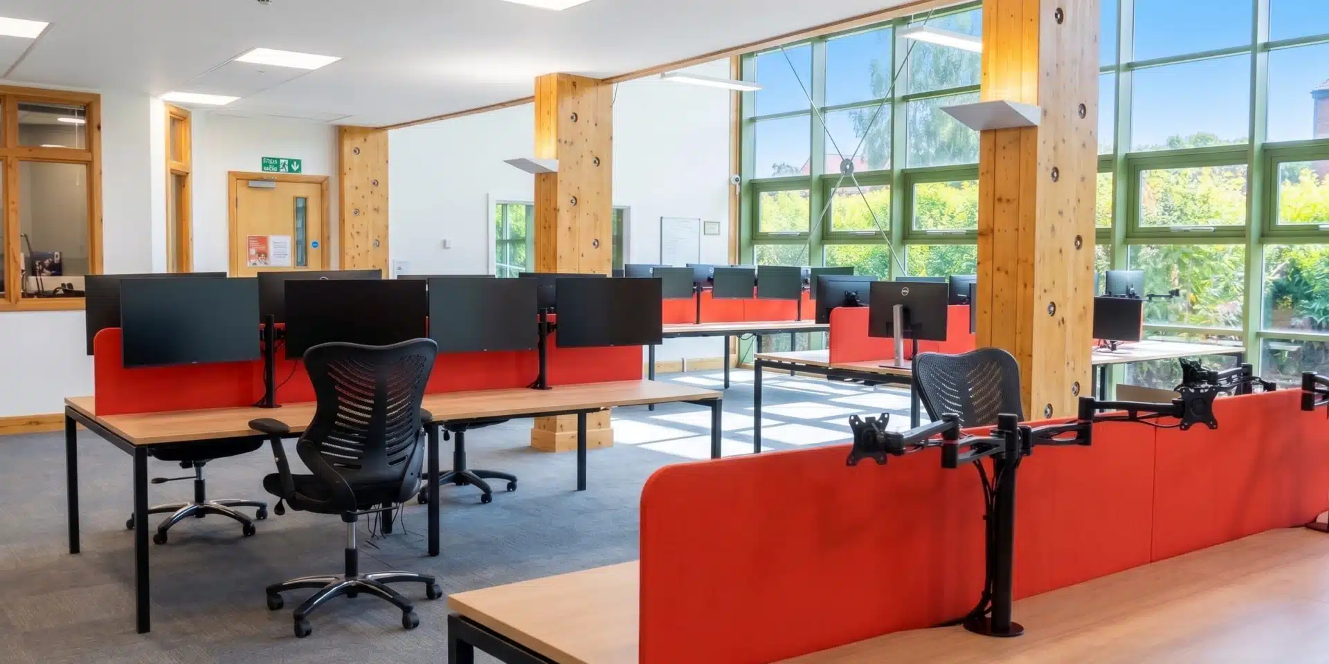 Empty office desks with red partition screens and monitor arms next to large windows.