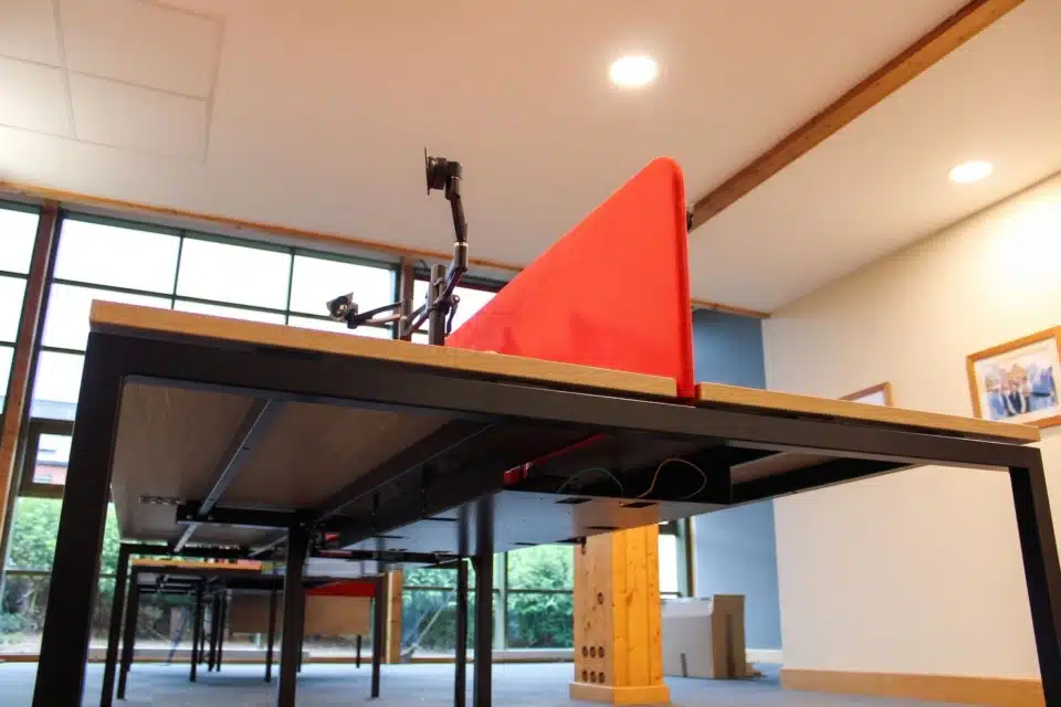 Empty office desks with red partition screens and monitor arms next to large windows.