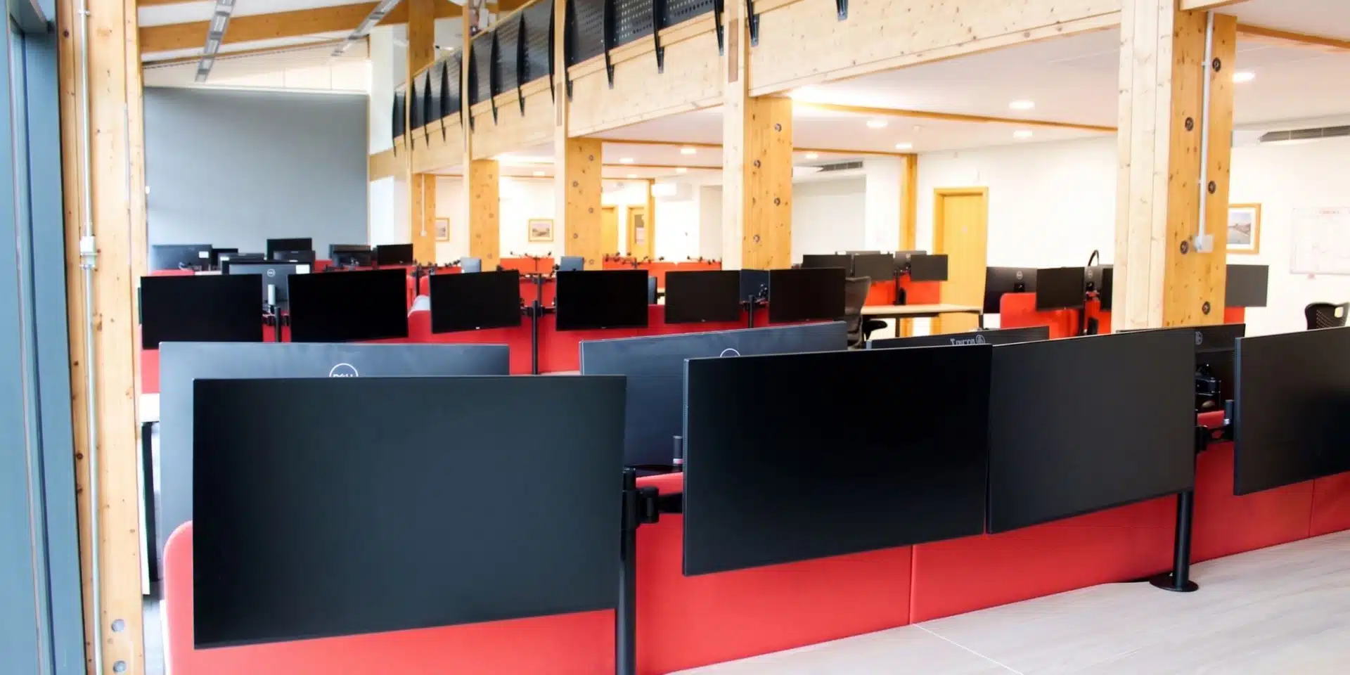 Sunlit office interior featuring modern workstations with red dividers and black monitor stands.