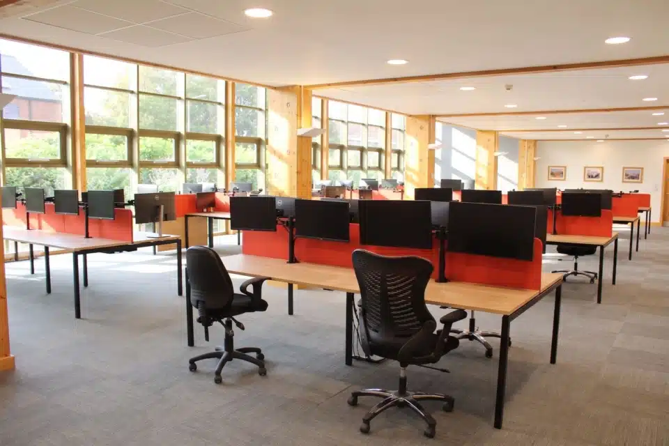Sunlit office interior featuring modern workstations with red dividers and black monitor stands.